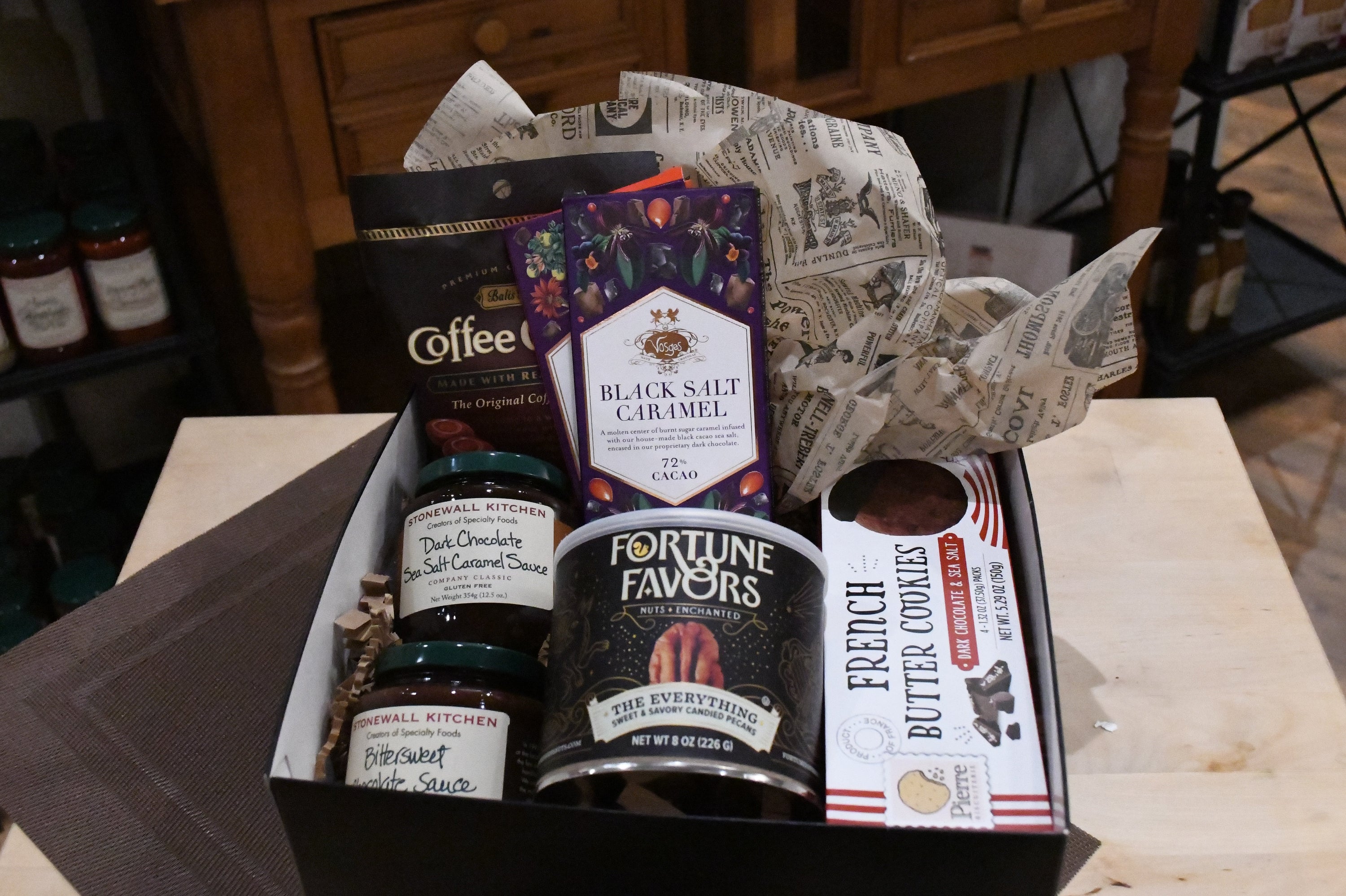 A curated gourmet gift box filled with assorted snacks and treats, including a bag of Coffee Town coffee, Fortune Favors chocolate, French Butter Cookies, and jars of preserves, arranged in a black box with newspaper-style wrapping paper.