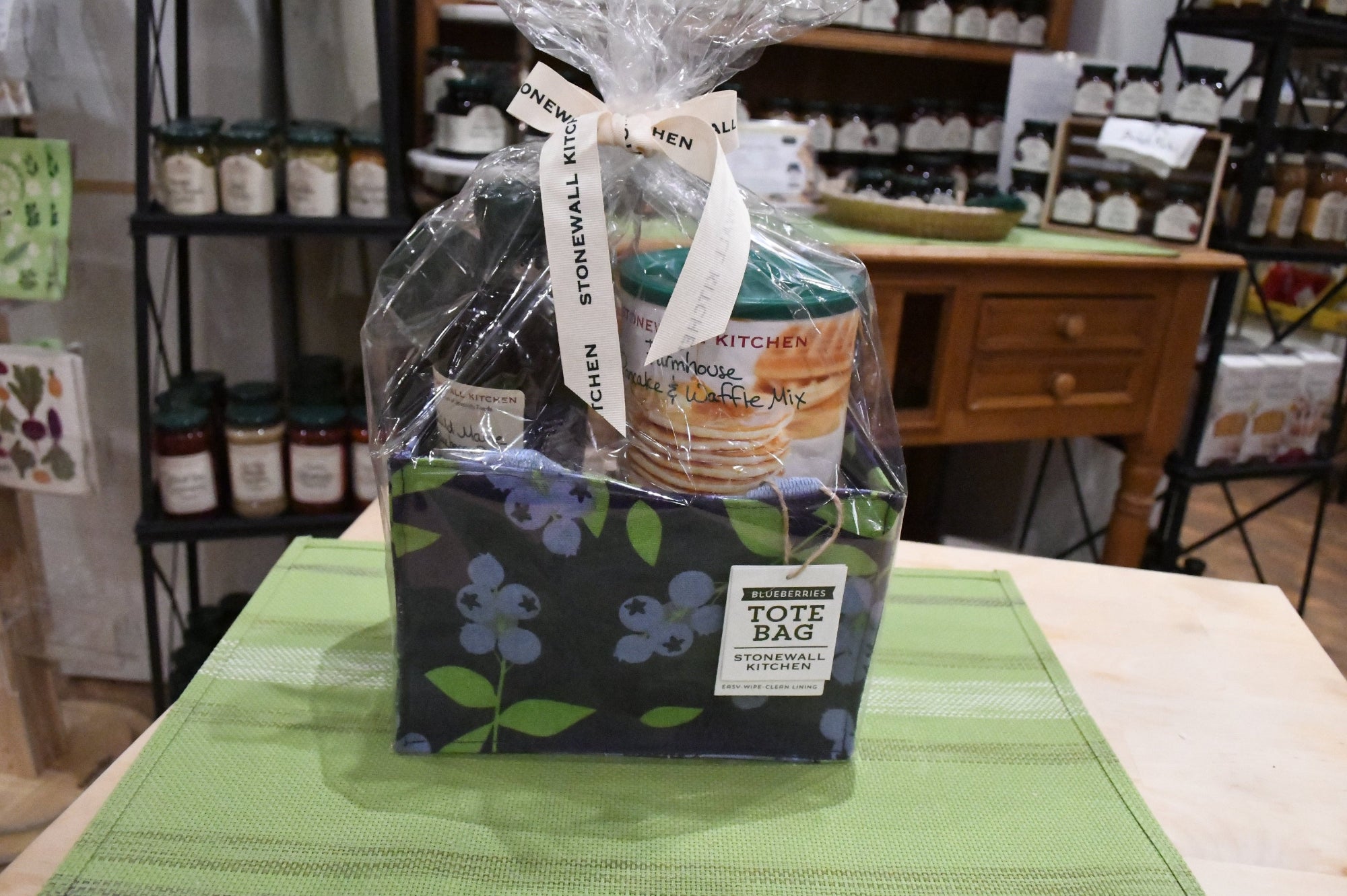A Storwall Kitchen Tote Bag gift set wrapped in clear cellophane and tied with a white ribbon, displayed on a green placemat in a retail store setting.