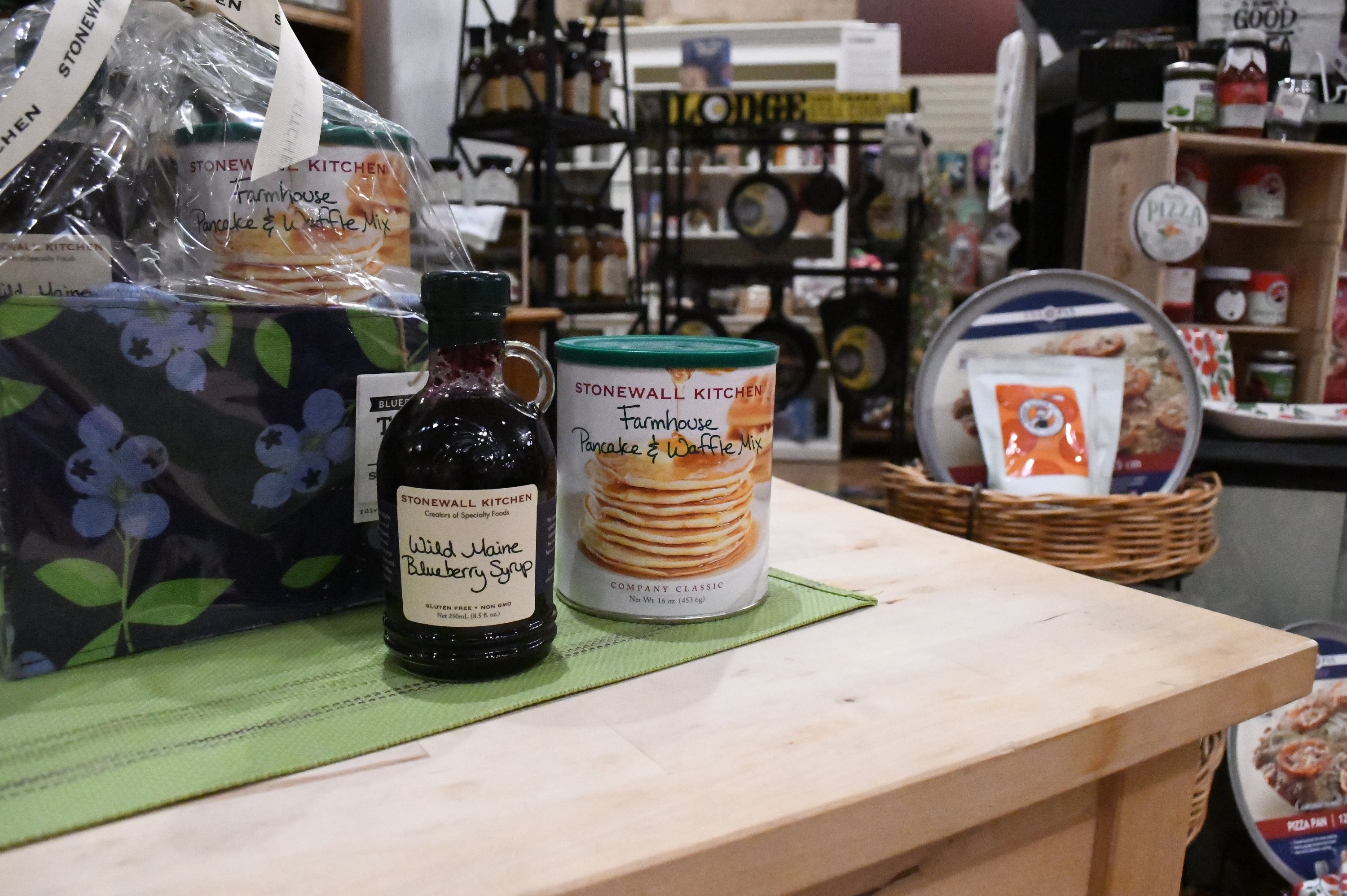 A countertop display in a rustic shop or lodge setting featuring a bottle of Stonewall Kitchen Blueberry Jam and a can of Stonewall Kitchen Frozen Treats, alongside a floral napkin and a basket of packaged goods.
