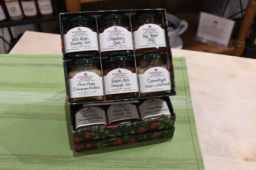 A boxed assortment of small glass jars of spreads and preserves arranged in a black gift tray on a green striped cloth. The labels show flavors including Wild Almond Plumage Jam, Strawberry Spread, Hot Apple Syrup, Handmade Champagne Muffins, Apple Butter Chopped Apple, Caramelized Cream Cheese, and more.