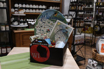 A gift basket box filled with a large Michigan-themed plate, a printed booklet, and small packaged items, displayed on a table inside a shop with shelves of jars and products in the background.