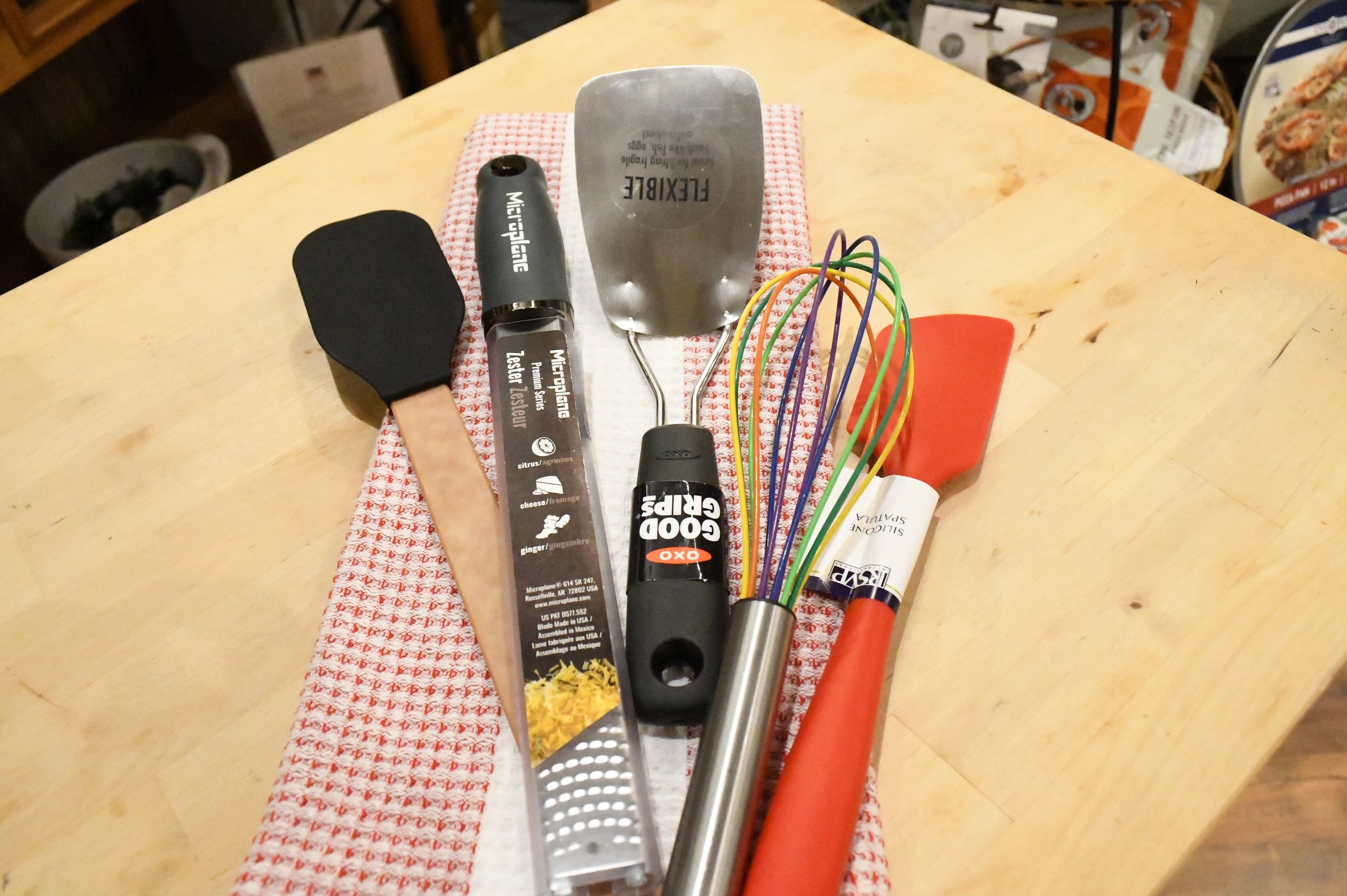 A set of kitchen utensils including a black spatula, metal spatula, whisk, and orange spatula resting on a red-and-white checkered towel on a light wood table.