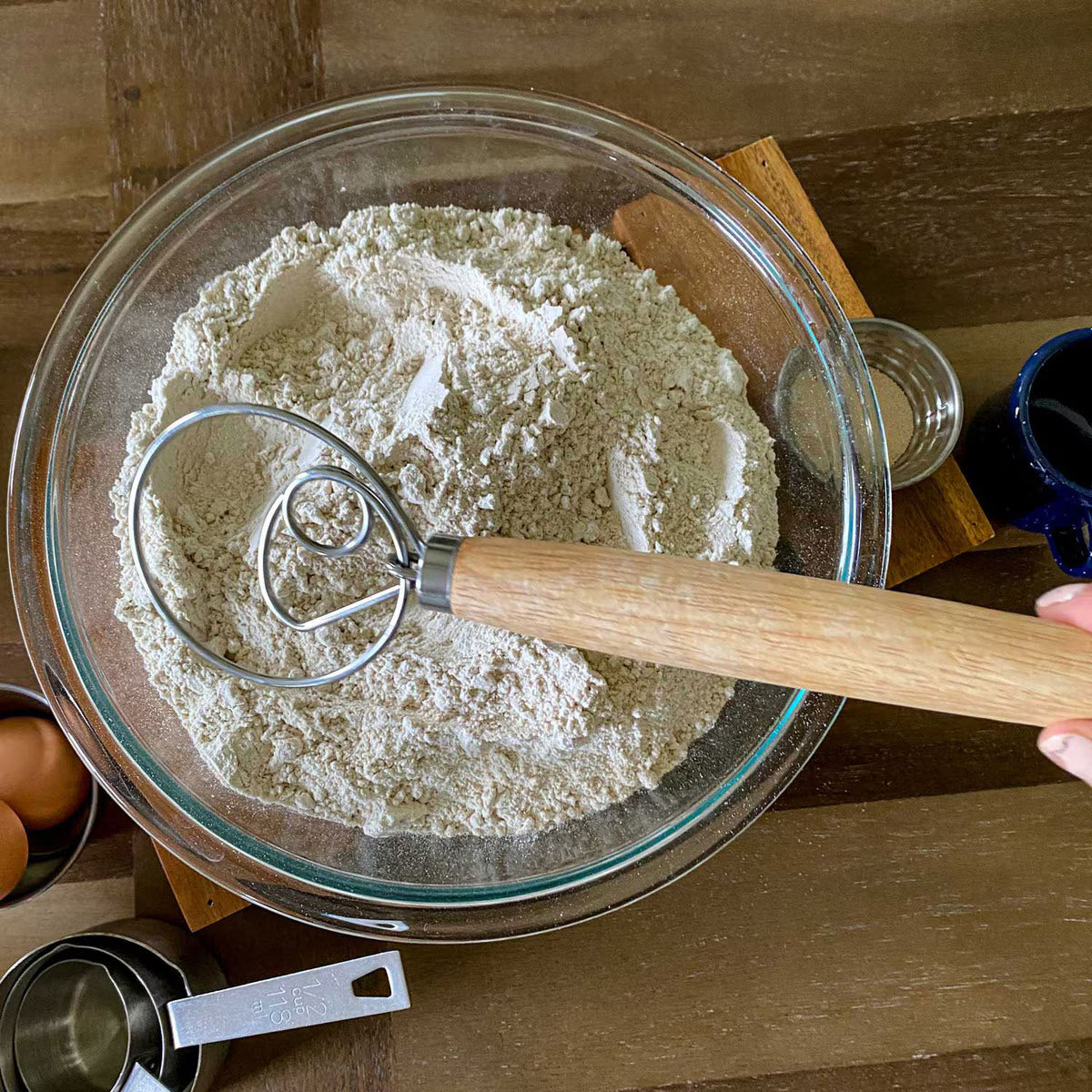 Glass bowl with flour and a Danish whisk on a wooden surface