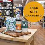 Table with a mug, gift box, and bottle of ginger ale with 'Free Gift Wrapping' text in a store setting.