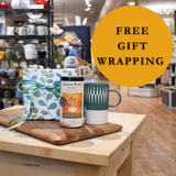 Table with a mug, gift box, and bottle of ginger ale with 'Free Gift Wrapping' text in a store setting.
