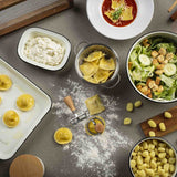 Tabletop scene with various pasta dishes and ingredients on a gray surface.