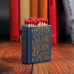 Matchbox labeled 'Slow Burn' with matches on a wooden surface, books in the background
