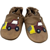 A pair of brown suede baby booties featuring a colorful patchwork fire truck applique on the side, resting on a rustic wooden surface against a white wood plank background.