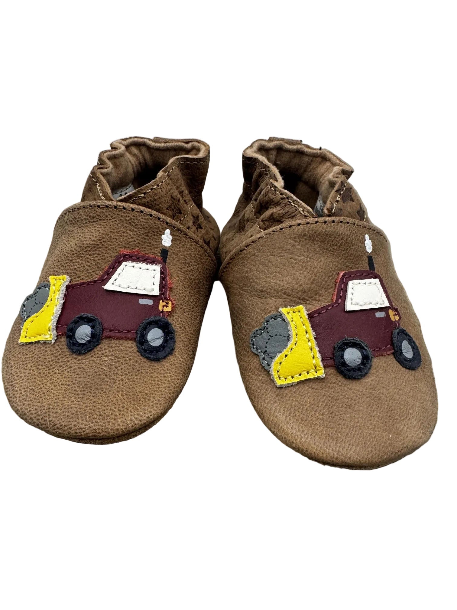 A pair of brown suede baby booties featuring a colorful patchwork fire truck applique on the side, resting on a rustic wooden surface against a white wood plank background.